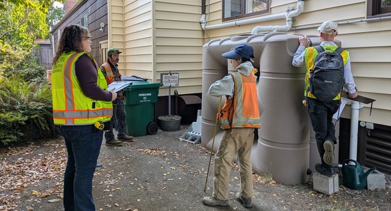 Four people in hi-vis vests look at a cistern next to a building. 