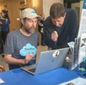 A man wearing a RainWise shirt and baseball cap sits at a laptop while another man looks over his shoulder