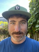 head shot photo of white man with a dark mustache wearing a King County baseball hat and royal blue t-shirt