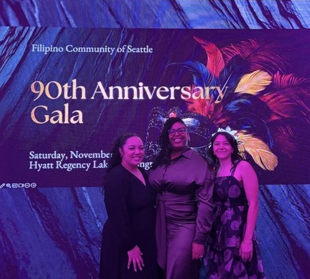 Three OPA staff standing in front of the 90th Anniversary Gala Sign. They are wearing formal dresses and smiling.