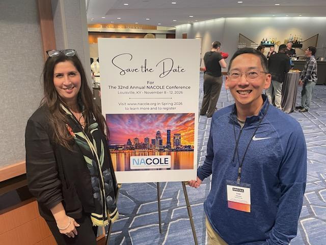 OPA Staff standing in the lobby of a conference center in front of the Save the Date poster. They are dressed in business casual and smiling.