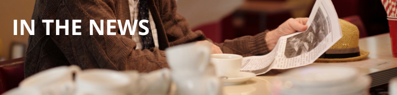 Photo of a gentleman reading a newspaper at a diner counter. There are white coffee mugs stacked up next to him.