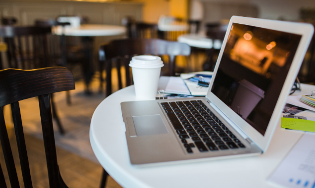 Photo of a laptop sitting open on a table at a cafe. There is a disposable coffee cup with a lid next to it, and papers scattered around.
