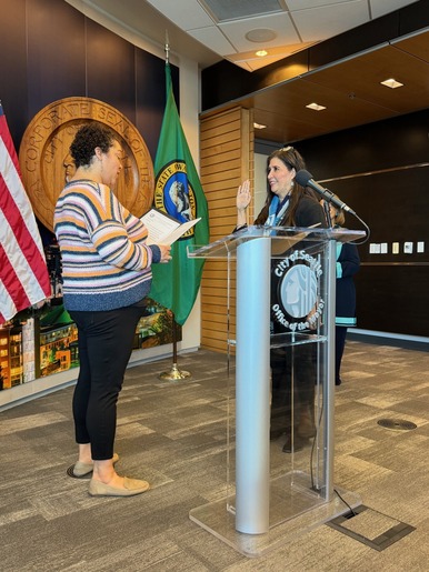 Bonnie Glenn being sworn in at City Hall by the City Clerk. Her right hand is up. There is a Washington State flag and American Flag behind her.
