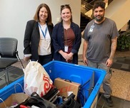 Three people stand beside a blue bin, with a laptop and various items placed on top of it.