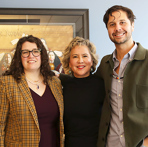 A woman with long, curly brown hair and glasses, a woman with short light hair, and a tall man with short brown hair stand together in an office.