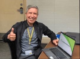 A man sitting at a table with a laptop, smiling and giving a thumbs up.