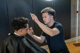 A barber with short black hair gives a haircut to a youth in a gymnasium