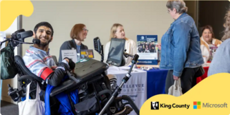 King County resource fair: Man in a wheelchair, smiling, with people standing by display table. Microsoft & King County logo placed in corner.