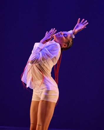 Photo of dancer in striking pose wearing white in front of an indigo background.
