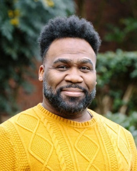 Photo of a poet with a black beard speckled with gray and kind eyes wearing a bright yellow sweater.