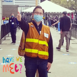 A Chinese man wearing a orange safety vest and blue medical mask with his arm extended giving a thumbs up