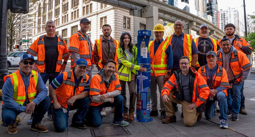 A group SPU employees pose next to a blue, SPU-branded water refill station in Downtown Seattle