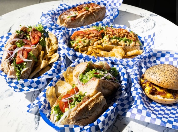 A photo of a colorful spread of sub sandwiches, chopped cheese, and chips on checkered blue and white wax paper.