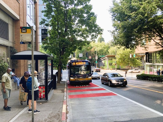 A King County Metro Route 8 bus approaches a stop on Westlake Avenue North in Seattle as several people wait beside a bus shelter.