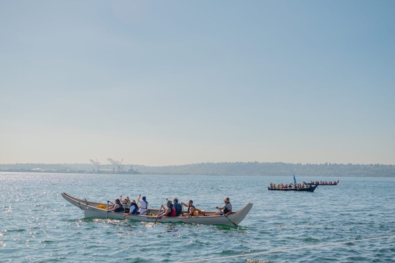 Three long canoes, filled with people rowing, on the water on a sunny day.