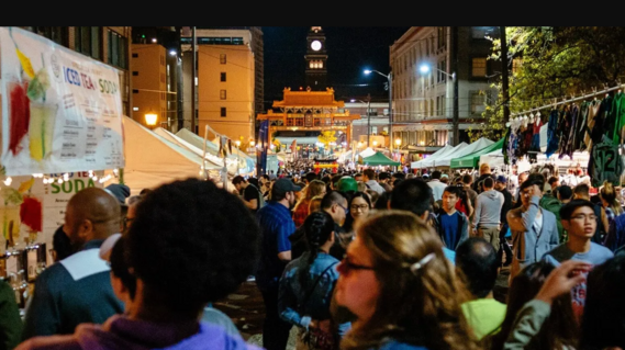 Large crowd browsing illuminated vendor stalls at a night market, with a traditional Chinese gate and clock tower in the background.