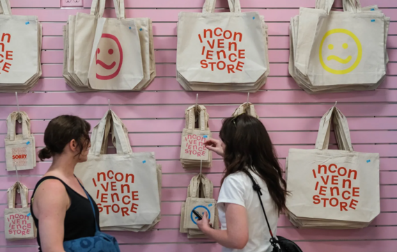 Two people browse a pink slatwall display of cream tote bags reading "Inconvenience Store" with slanted emoji faces and "SORRY" gift bags.