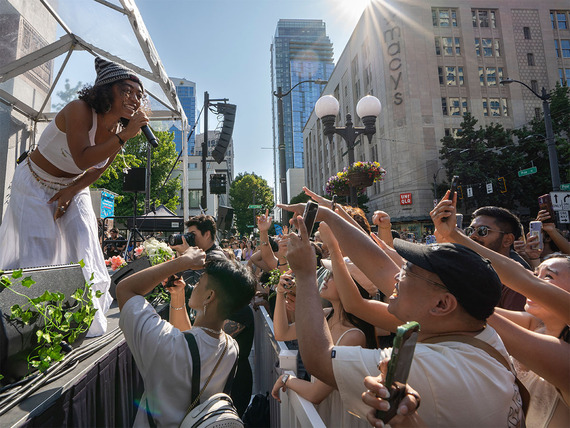 Singer in white outfit performs on an outdoor stage, smiling down at a crowd of fans in a sunny downtown street setting.