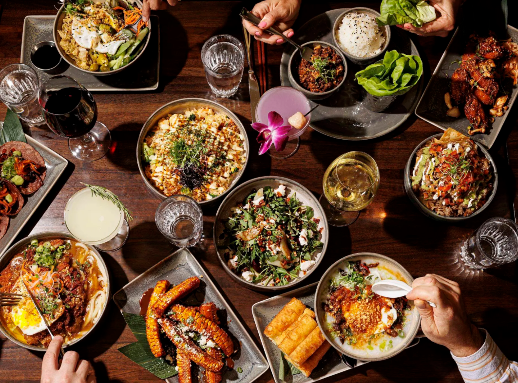 Overhead view of a dark wooden table filled with colorful Asian-fusion dishes, cocktails, and shared plates as diners reach in to eat together.