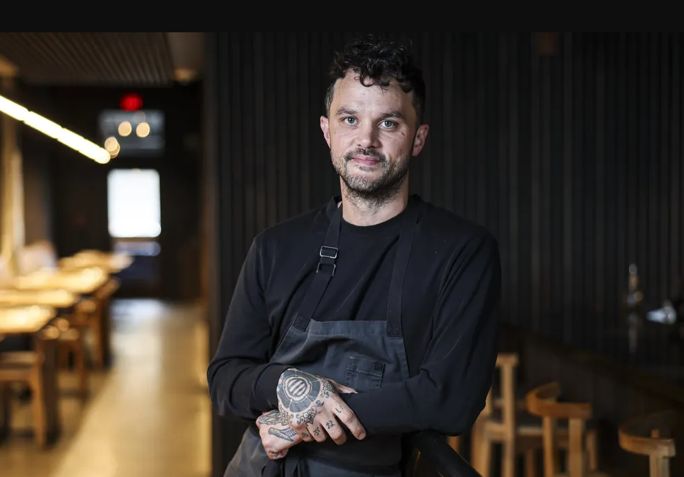 Brady Ishiwata Williams, a chef in a dark apron, leans forward with tattooed hands inside a dimly lit restaurant dining room.