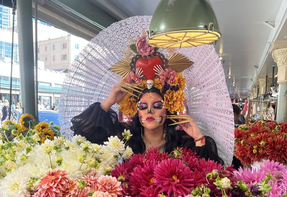 Person in Día de los Muertos makeup and floral headdress poses behind colorful dahlias at Pike Place Market.