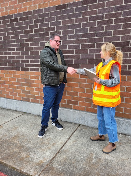 A man in plainclothes and a woman in an orange safety vest shake hands in front of a brick wall.