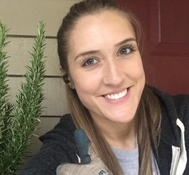 Close-up photo of Madelyn Loy (a young blonde woman) smiling, standing outside of a house next to a rosemary plant.