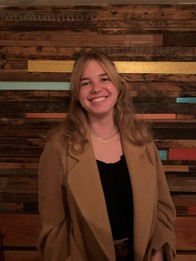 Photo of Anna Thomas, a young blonde woman in a black shirt and brown coat standing in front of a wood wall.