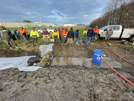A group of people in safety and work gear looking at a ditch full of water with a sandbag and plastic sheet barrier set up in it.