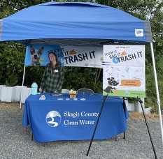 A person smiling under a pop-up tent. They are behind a table with a blue tablecloth reading "Skagit County Clean Water."