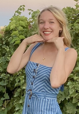 A young blonde woman in a blue dress in a vineyard at sunset. She is posing for the photo with a smile and her hands framing her face.