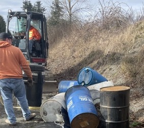 More 55-gallon barrels on the side of the road, with workers and a small excavator visible in the background.