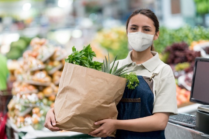 Masked grocery worker carries full brown paper grocery bag