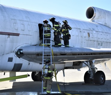 Three firefighters stand on wing of decommissioned 727 airplane while fourth holds ladder during emergency exercise in 2018