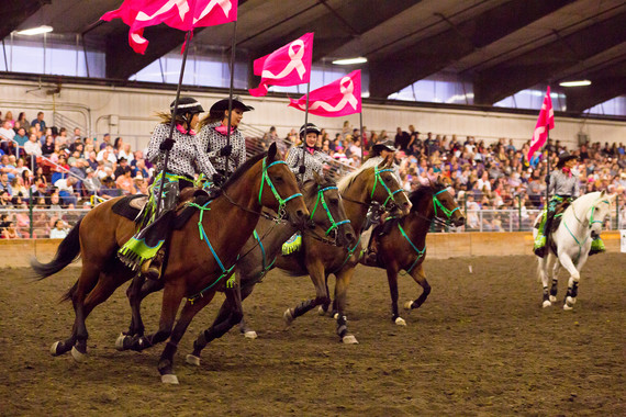 Horse riders with pink breast cancer awareness flags ride in an equestrian arena.