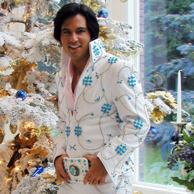 Danny Vernon dressed as Elvis Presley in a white and turquoise suit by a blue and white Christmas tree. 