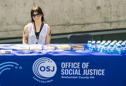 Office of Social Justice table with bubbles, books, stickers, literature about the OSJ, Administrative Assistant Cortney Murphy sitting at the table