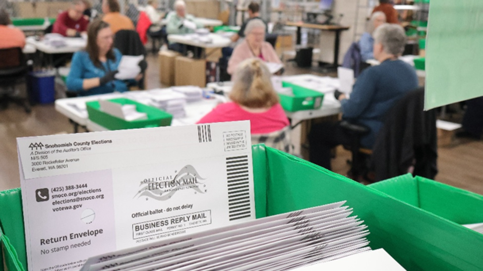 Batches of Snohomish County ballots sit in green trays awaiting processing, while election workers process ballots in the background.