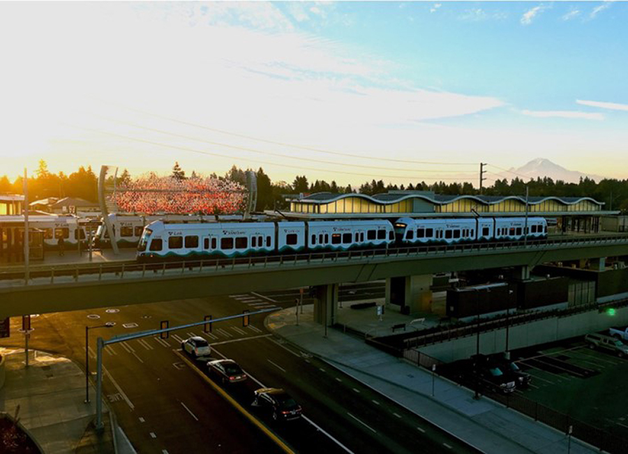 A photo of a Link light rail train crossing a bridge at sunset with the silhouette of Mt. Rainier in the background
