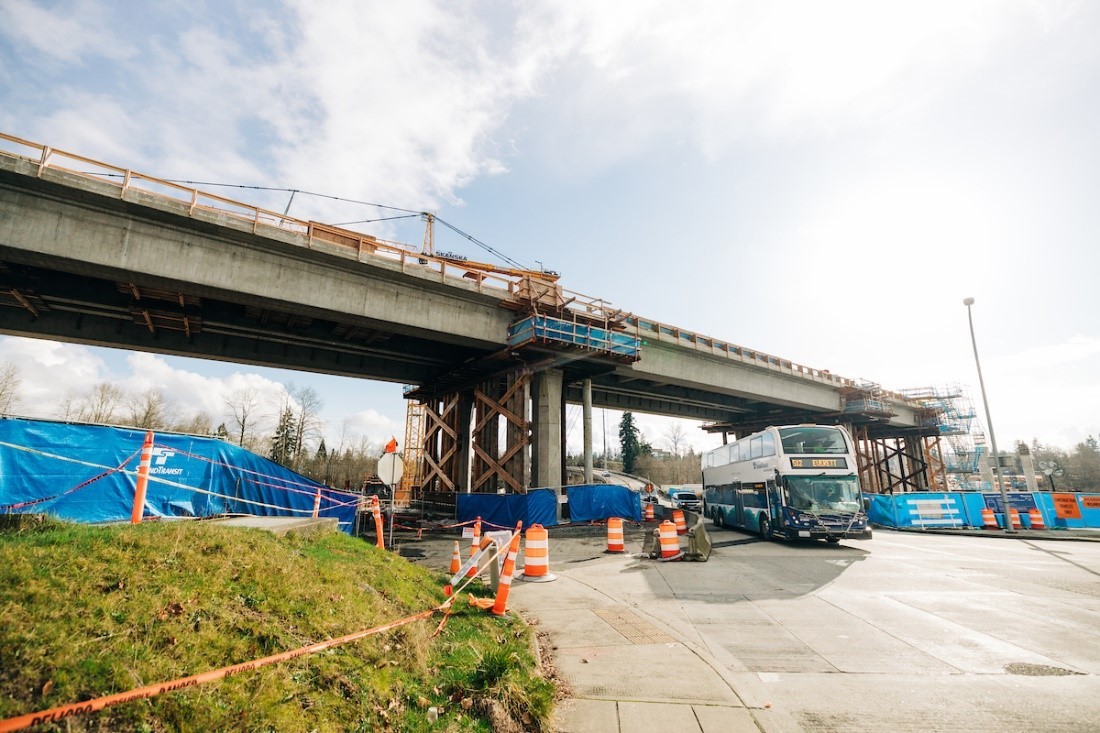 Photo of A Sound Transit double-decker bus driving under a light rail track span. 