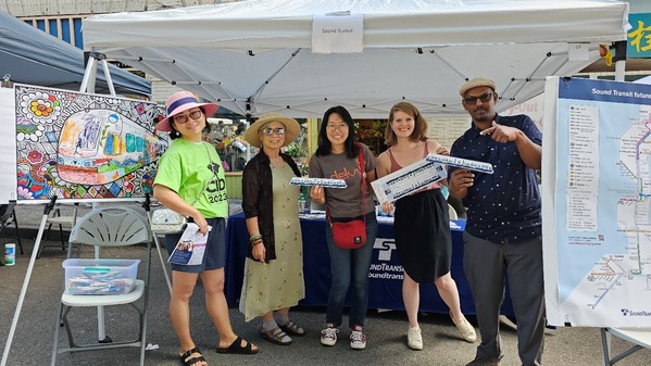 A group of community liaisons, Sound Transit staff, and outreach support staff, smiling and holding flyers and cardboard train cutouts at a event. 