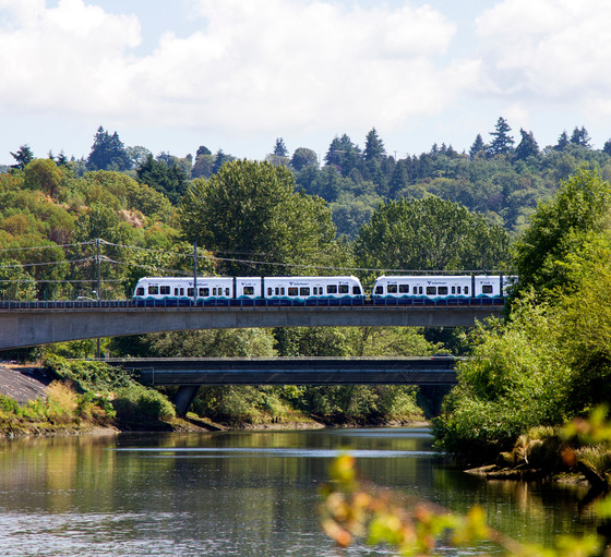 A link light rail train traveling over a river on a bridge surrounded by greenery on a sunny day.