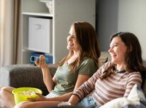 Two smiling women sitting together on a couch, one holding a bucket of popcorn and a remote control.