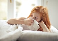 Young girl holding tissue to her nose laying under blankets in a bed.