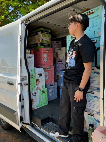 Young man standing in the side doorway of a van packed with boxes of diapers.