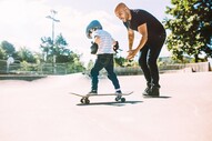 Young boy skateboard with dad looking on, encouragingly.