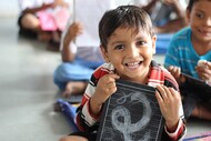Smiling boy holding a chalkboard in the foreground and other children, blurred, in the background.