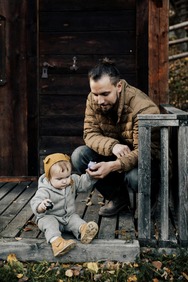 Man crouched holding the hand of a baby who is sitting on a porch in a fall setting.