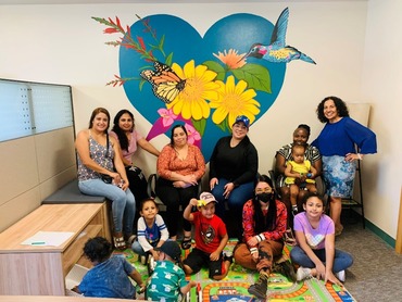 A group of people gathered in front of a mural of flowers, a hummingbird, and butterfly overlaying a blue heart.